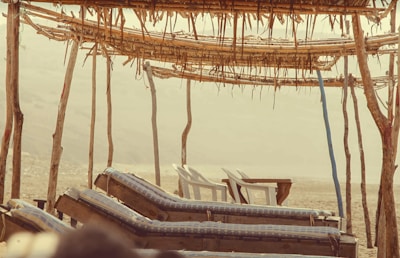 A rustic beach setting with wooden lounge chairs covered in blue checkered cushions. The loungers face the open landscape, sheltered by a simple canopy made from poles and thatched materials. Two white plastic chairs and a table are visible in the background, suggesting a relaxed, low-key atmosphere.