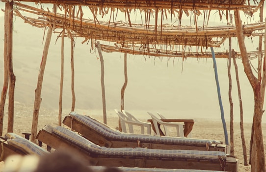 A rustic beach setting with wooden lounge chairs covered in blue checkered cushions. The loungers face the open landscape, sheltered by a simple canopy made from poles and thatched materials. Two white plastic chairs and a table are visible in the background, suggesting a relaxed, low-key atmosphere.