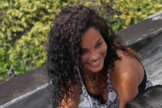 A person with curly hair smiles while leaning on a wooden surface with greenery in the background.