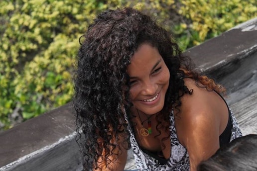 A person with curly hair smiles while leaning on a wooden surface with greenery in the background.
