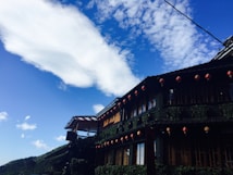 An elegant, traditional-style building with multiple floors is adorned with red lanterns and lush green foliage. The blue sky above features a striking formation of white clouds, and the structure is nestled against a backdrop of verdant hills.
