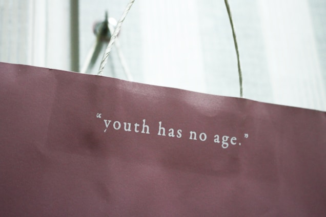 A maroon paper bag with a white rope handle features the quote 'youth has no age' printed in white lettering.