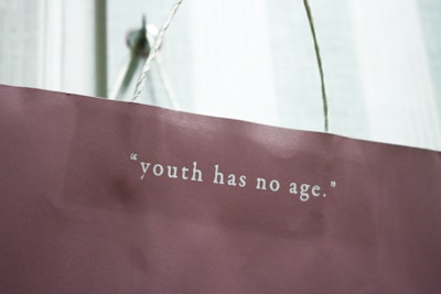 A maroon paper bag with a white rope handle features the quote 'youth has no age' printed in white lettering.