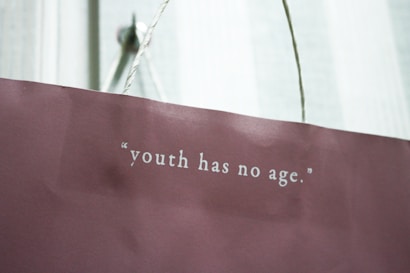 A maroon paper bag with a white rope handle features the quote 'youth has no age' printed in white lettering.
