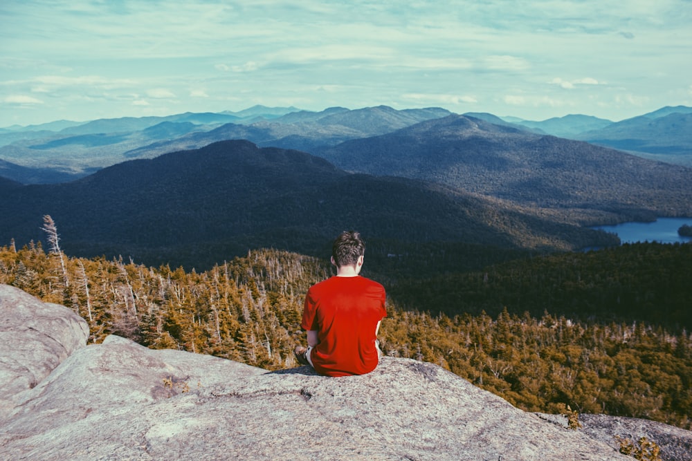 man sitting on gray rock formation looking at pine trees during daytime