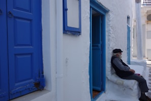 An elderly person wearing a black hat and jacket sits on a white cushion against a whitewashed building with vibrant blue doors and windows. The setting appears bright and sunlit, typical of a Mediterranean locale.