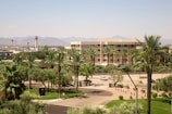 A landscaped urban area featuring a modern hotel or office building surrounded by numerous palm trees and greenery. In the background, a desert scene with distant mountains and a control tower rising above a cityscape.