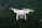 A white drone with a camera is flying against a blurred background of dense green forest. The propellers are visible as they spin, indicating motion. The drone appears to be hovering steadily.