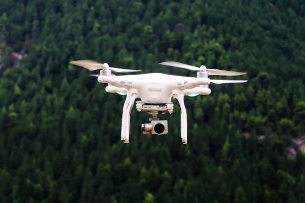 A white drone with a camera is flying against a blurred background of dense green forest. The propellers are visible as they spin, indicating motion. The drone appears to be hovering steadily.