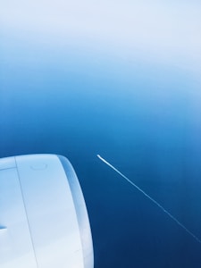 A section of an airplane wing and engine is visible against a vast expanse of blue sky. There is a thin white contrail stretching diagonally through the sky, suggesting the presence of another aircraft in the distance.