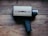 A vintage Super 8 movie camera sits on a wooden surface. The camera has a tan body with a label reading 'argus 806' and 'super eight.' It features a black handle and a textured focus ring on the lens.