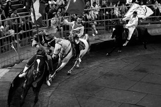 Three riders on horseback are engaged in what appears to be a dynamic, fast-paced equestrian performance or game. They are in traditional attire and holding flags while racing around a fenced arena. Spectators in the background watch the action with interest.