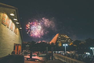 Fireworks light up the night sky above a modern pyramid-shaped building and a crowded pier. The foreground features a wooden building with 'EAT A LOT' written on its side, while the water reflects the lights from the scene. The atmosphere is lively, with people gathered to watch the display.