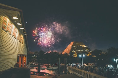 Fireworks light up the night sky above a modern pyramid-shaped building and a crowded pier. The foreground features a wooden building with 'EAT A LOT' written on its side, while the water reflects the lights from the scene. The atmosphere is lively, with people gathered to watch the display.