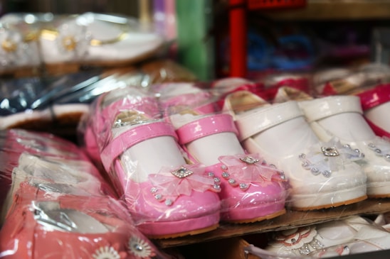 Brightly colored children's shoes are displayed closely together. The shoes are made of glossy materials and come in shades of pink and white. Some of the shoes feature decorative accents such as floral designs and small bows.