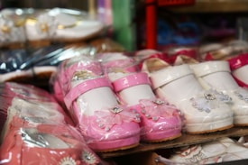 Brightly colored children's shoes are displayed closely together. The shoes are made of glossy materials and come in shades of pink and white. Some of the shoes feature decorative accents such as floral designs and small bows.