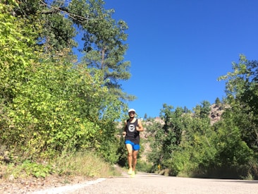 A person is jogging along a paved path surrounded by lush green trees under a clear blue sky. The individual is wearing a white cap, a black tank top, blue shorts, and bright running shoes. The setting appears to be a scenic outdoor area with hills covered in trees.