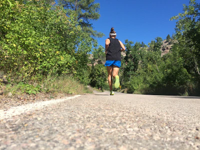 Active runner tying shoes on a sunny trail surrounded by green trees and clear skies.