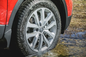 A close-up view of a car wheel with a red fender above it, partially submerged in shallow water. The tire is new and clean, with visible treads, and the reflection of the rim is seen on the wet surface.