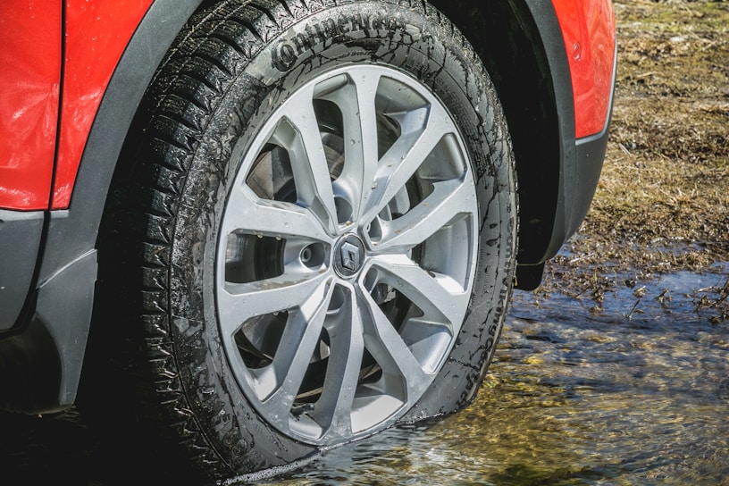 A close-up view of a car wheel with a red fender above it, partially submerged in shallow water. The tire is new and clean, with visible treads, and the reflection of the rim is seen on the wet surface.
