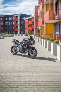 A modern black motorcycle is parked on a paved courtyard in front of a contemporary apartment building. The building features a striking design with red railings and yellow accents, complemented by neatly arranged greenery in large, white planters. The sky is partly cloudy, providing a bright and inviting atmosphere.