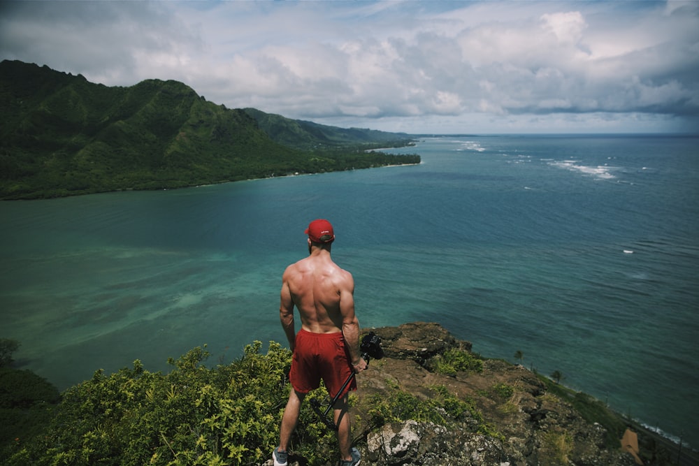Hombre en topless en pantalones cortos rojos de pie en un acantilado de roca