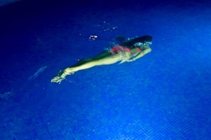 A person is swimming underwater in a pool, wearing a pink swimsuit. The pool's tiles are blue, and the water is clear, with gentle ripples created by the swimmer's movement.
