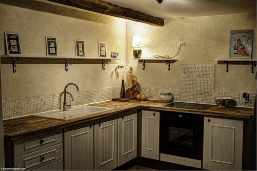 A cozy kitchen corner featuring wooden cabinetry and a countertop with various kitchen items. Shelves mounted on the wall hold decorative pieces, including framed pictures and a small lamp. The lighting creates a warm and inviting atmosphere.