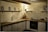 A cozy kitchen corner featuring wooden cabinetry and a countertop with various kitchen items. Shelves mounted on the wall hold decorative pieces, including framed pictures and a small lamp. The lighting creates a warm and inviting atmosphere.