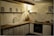 A cozy kitchen corner featuring wooden cabinetry and a countertop with various kitchen items. Shelves mounted on the wall hold decorative pieces, including framed pictures and a small lamp. The lighting creates a warm and inviting atmosphere.
