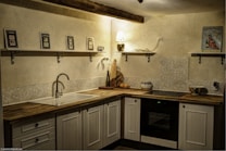 A cozy kitchen corner featuring wooden cabinetry and a countertop with various kitchen items. Shelves mounted on the wall hold decorative pieces, including framed pictures and a small lamp. The lighting creates a warm and inviting atmosphere.