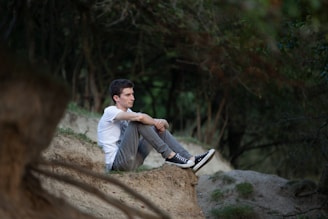 A person sits on a sandy embankment in a forested area, with legs pulled close to the chest. The setting is serene, with a backdrop of trees and natural greenery. The individual appears to be deep in thought or contemplation.