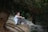 A person sits on a sandy embankment in a forested area, with legs pulled close to the chest. The setting is serene, with a backdrop of trees and natural greenery. The individual appears to be deep in thought or contemplation.