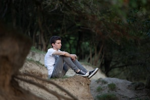 A person sits on a sandy embankment in a forested area, with legs pulled close to the chest. The setting is serene, with a backdrop of trees and natural greenery. The individual appears to be deep in thought or contemplation.