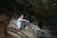 A person sits on a sandy embankment in a forested area, with legs pulled close to the chest. The setting is serene, with a backdrop of trees and natural greenery. The individual appears to be deep in thought or contemplation.