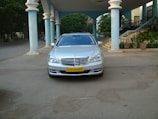 A silver luxury sedan is parked under a grand, covered entrance with tall pillars. The car has a clean, polished look and an identifiable yellow license plate. Surrounding the entrance are plants and stairs leading up to a building.