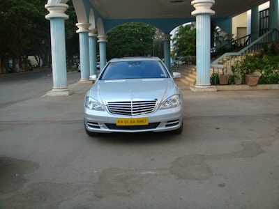 A silver luxury sedan is parked under a grand, covered entrance with tall pillars. The car has a clean, polished look and an identifiable yellow license plate. Surrounding the entrance are plants and stairs leading up to a building.