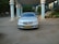 A silver luxury sedan is parked under a grand, covered entrance with tall pillars. The car has a clean, polished look and an identifiable yellow license plate. Surrounding the entrance are plants and stairs leading up to a building.