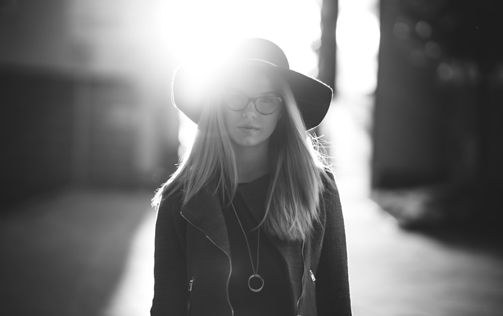 grayscale photography of woman wearing hat and coat standing near wall