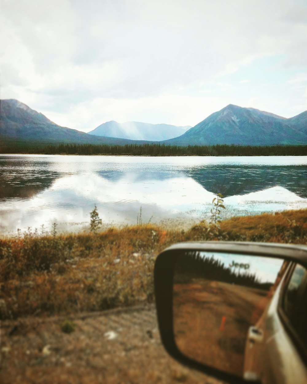 depth photography of car side mirror with a scene of body of water near mountains