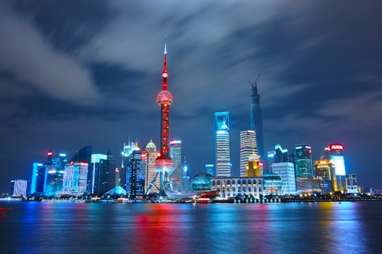 A vibrant and illuminated cityscape of Shanghai at night with the Oriental Pearl Tower prominently featured among various skyscrapers reflecting brightly on the river. The buildings display a mix of modern architecture, with colorful lights creating a lively atmosphere.