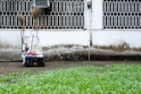 A cleaning cart is positioned against a wall with a geometric metal grille window above. It contains brooms and a mop. The wall shows signs of weathering, with a distinct dirty line. Green grass grows in the foreground.
