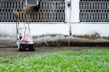 A cleaning cart is positioned against a wall with a geometric metal grille window above. It contains brooms and a mop. The wall shows signs of weathering, with a distinct dirty line. Green grass grows in the foreground.