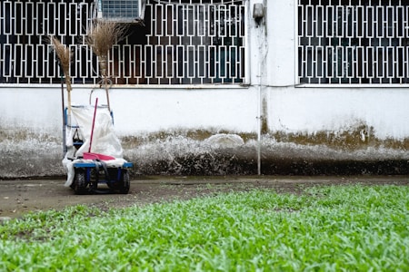 A cleaning cart is positioned against a wall with a geometric metal grille window above. It contains brooms and a mop. The wall shows signs of weathering, with a distinct dirty line. Green grass grows in the foreground.