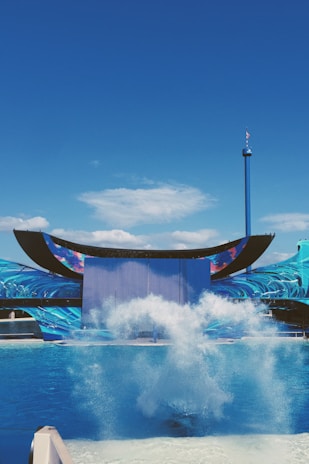 A dramatic splash occurs in a large blue pool set against a backdrop of an aquatic-themed stage with intricate wave designs. The sky is clear and deeply blue, and a tall tower with a flag can be seen in the background.