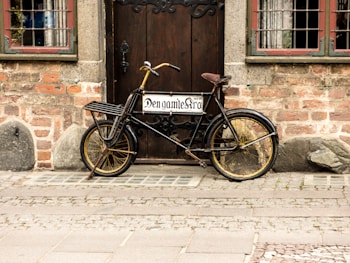 An old-fashioned black bicycle with a wooden cargo rack is parked against a rustic brick wall in front of a wooden door. The door features an ornate metal handle, and there are vintage-style windows on either side, with metal grid patterns and visible curtains. The scene conveys a nostalgic, historical atmosphere.