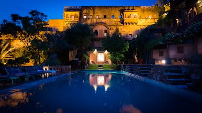 An illuminated historic building features prominently in the background, with intricate architecture and warm lighting that highlights its details. In the foreground, a tranquil pool reflects the structure, framed by lush greenery and surrounded by lounge chairs. The scene exudes a serene and luxurious ambiance, with a deep blue evening sky casting a peaceful glow.