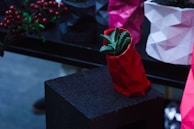 A geometric, faceted planter in bright red holds a green succulent plant. It is placed on a dark, rough-textured surface. In the background, several other planters are visible in varying colors such as pink, white, and black. Red berries are grouped at the edge of the scene, adding a pop of natural color.