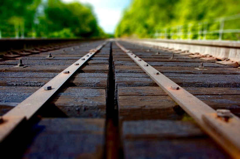 A close-up view of railway tracks, focusing on the wooden sleepers and metal rails. The image extends into a blurred background, surrounded by lush green trees. The perspective gives a sense of depth and distance.