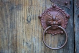 An intricately designed metal door knocker in the shape of an ornate face with a ring, mounted on a wooden door with visible grain texture.